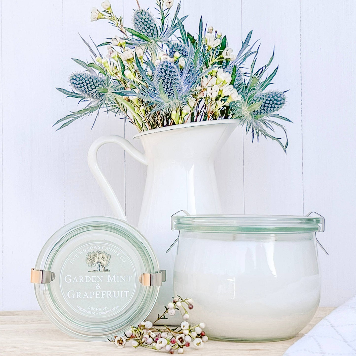 Decorative pitcher with flowers and two glass containers on a wooden surface.